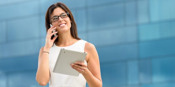 Business woman holding tablet pc computer and calling on cell phone outside corporate building. Caucasian hispanic female executive doing her job using digital communication technology.