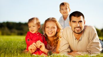 Happy family of 4 people lying ona grass under summer sun. Focus is on the man.