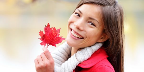 Fall girl holding red Autumn leave outside. Asian woman outdoor portrait in red seasonal autumn coat by fall forest lake. Female model smiling happy looking at camera. Mixed race Asian Caucasian girl.